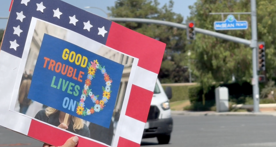 A person holds a sign at a street intersection with the phrase “GOOD TROUBLE LIVES ON” written in colorful letters next to a peace sign made of flowers. The sign is bordered in red, white, and blue with stars, resembling the American flag. In the background, a street sign reads “McBean Pkwy” and a white van waits at a red light.